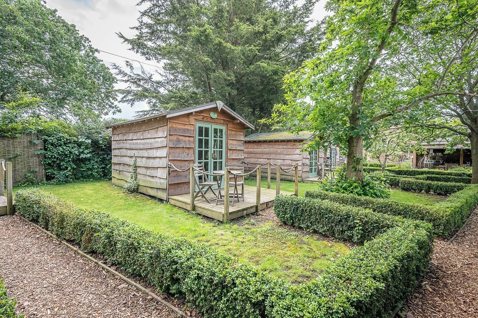 Una cabaña de madera con puerta verde, rodeada por un jardín cuidado con setos y una terraza de madera.