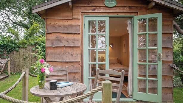 Une cabane en bois rustique avec une porte verte, une table ronde en bois entourée de chaises et un vase rempli de fleurs.