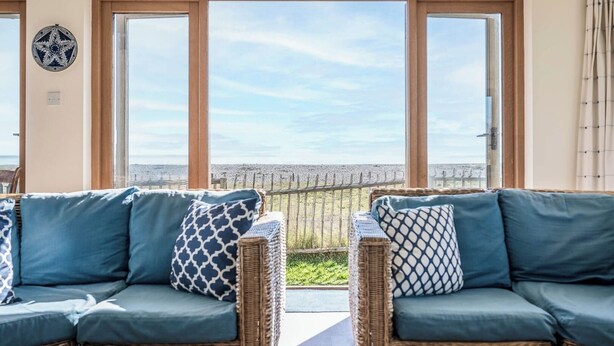 A room with two wicker sofas with blue cushions, a large window with a view of the beach, and a star-shaped decoration on the wall. - Fairhaven, Thorpeness (Thorpeness)