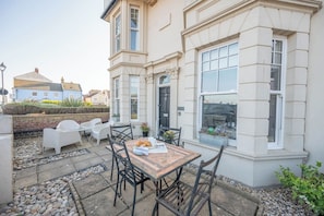 Une terrasse avec une table et des chaises, un petit jardin et un bâtiment doté de grandes fenêtres.