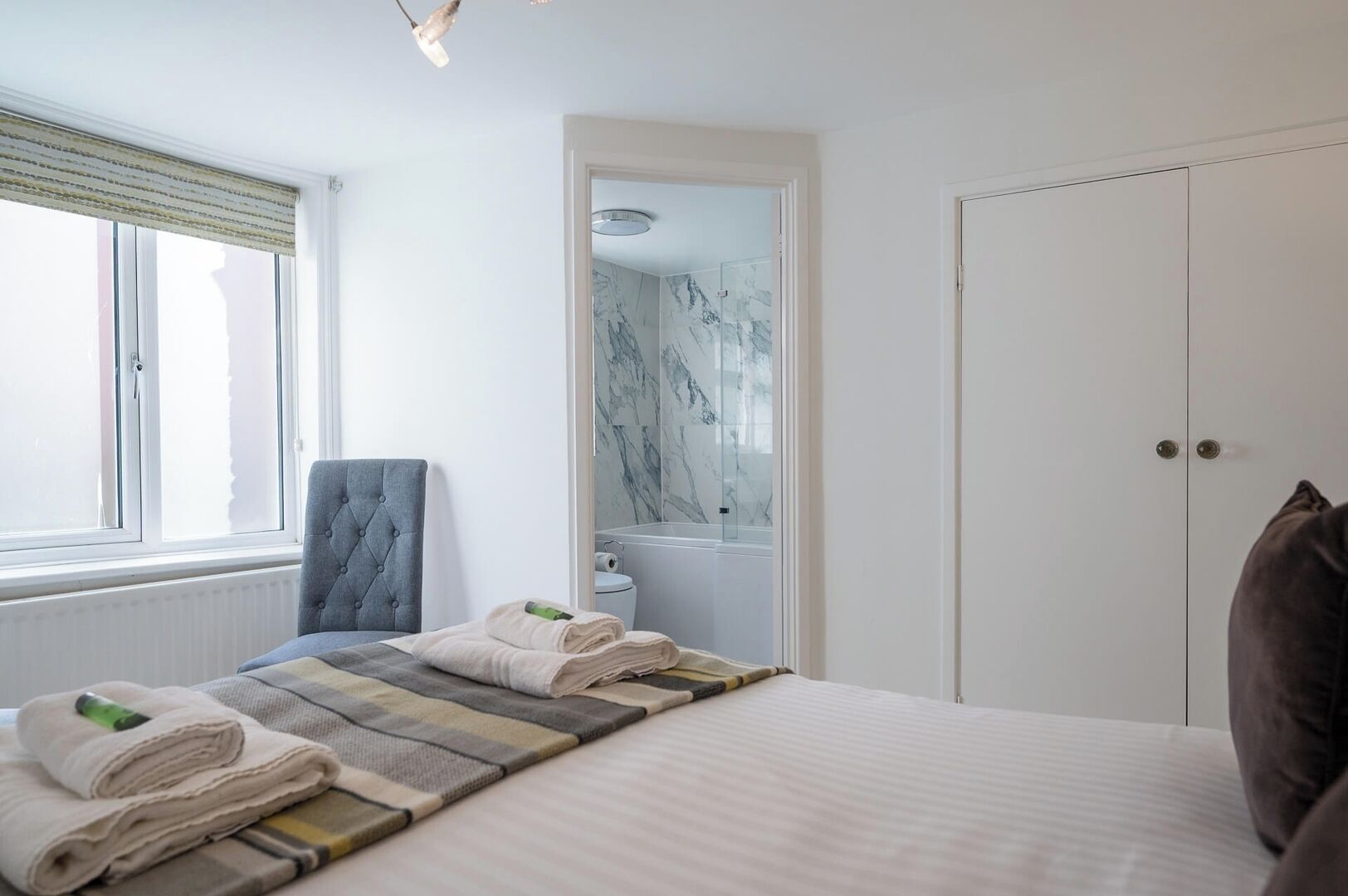 Une chambre à coucher avec un lit, une chaise, une fenêtre, une porte et une salle de bain visible à travers une porte ouverte.