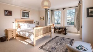 A bedroom with a wooden bed, a bedside table, a nightstand, a large window with curtains, and a floor lamp.