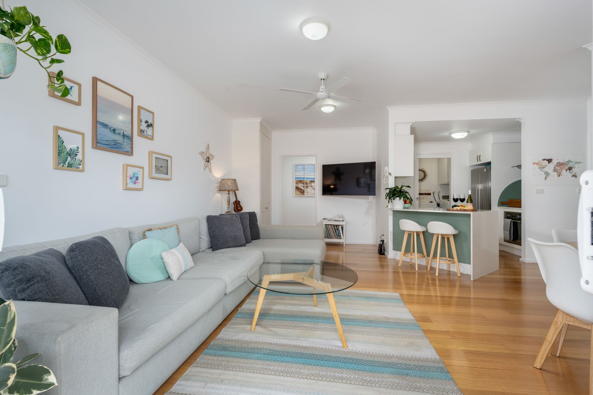 A modern living room with a grey sofa, a glass coffee table, and a striped rug. There is a kitchen area with white cabinets and a bar counter.