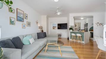 A modern living room with a grey sofa, a glass coffee table, and a striped rug. There is a kitchen area with white cabinets and a bar counter.