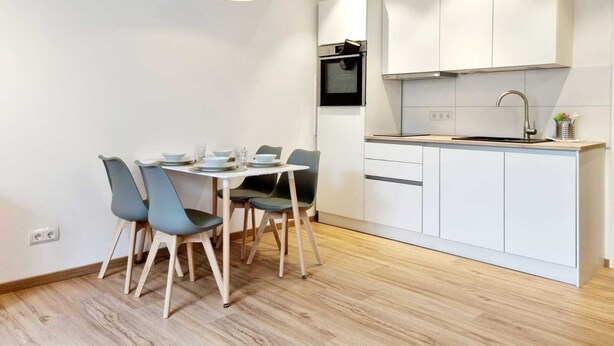 A modern kitchen with white cabinets, a built-in oven, and a dining area with a table and chairs. - Basement Apartment in the Munich/Ebersberg District—Affordable during the winter months—minimum stay of 7 days (Bruck)