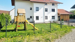 A multi-story building with a playground, including a slide and a wooden climbing structure, surrounded by a wire fence and greenery. - Basement Apartment in the Munich/Ebersberg District—Affordable during the winter months—minimum stay of 7 days (Bruck)