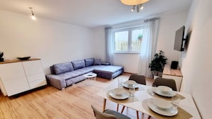 A modern living room with a dining area, a sofa, a TV, and a window with curtains. - Basement Apartment in the Munich/Ebersberg District—Affordable during the winter months—minimum stay of 7 days (Bruck)