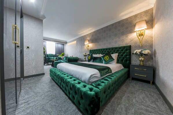 Un dormitorio amplio con una cama grande, un cabecero tapizado en verde y un banco a juego. Hay dos mesitas de noche con lámparas, un sofá verde y vistas al exterior a través de un ventanal.