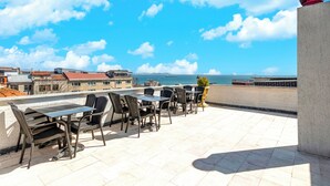 Una terrazza esterna con tavoli e sedie, vista sul mare e edifici sullo sfondo.