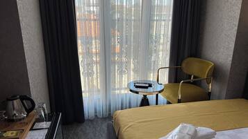 Ein Hotelzimmer mit Bett, einem kleinen runden Tisch mit Fernbedienung, einem Sessel, einem Schreibtisch mit Wasserkocher, Gläsern und einer Ablage mit Taschentüchtern.