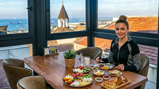 Un tavolo da pranzo con diversi cibi, una finestra che si affaccia su una località costiera e una persona seduta al tavolo.