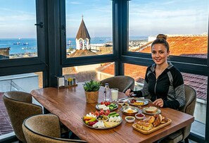 Una mesa de comedor con diversos alimentos, una ventana que ofrece vistas a un pueblo costero y una persona sentada a la mesa.