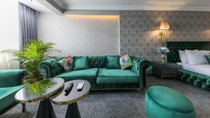 Una stanza con un divano verde, un tavolino rotondo, un letto con testiera verde e un comodino con una lampada.
