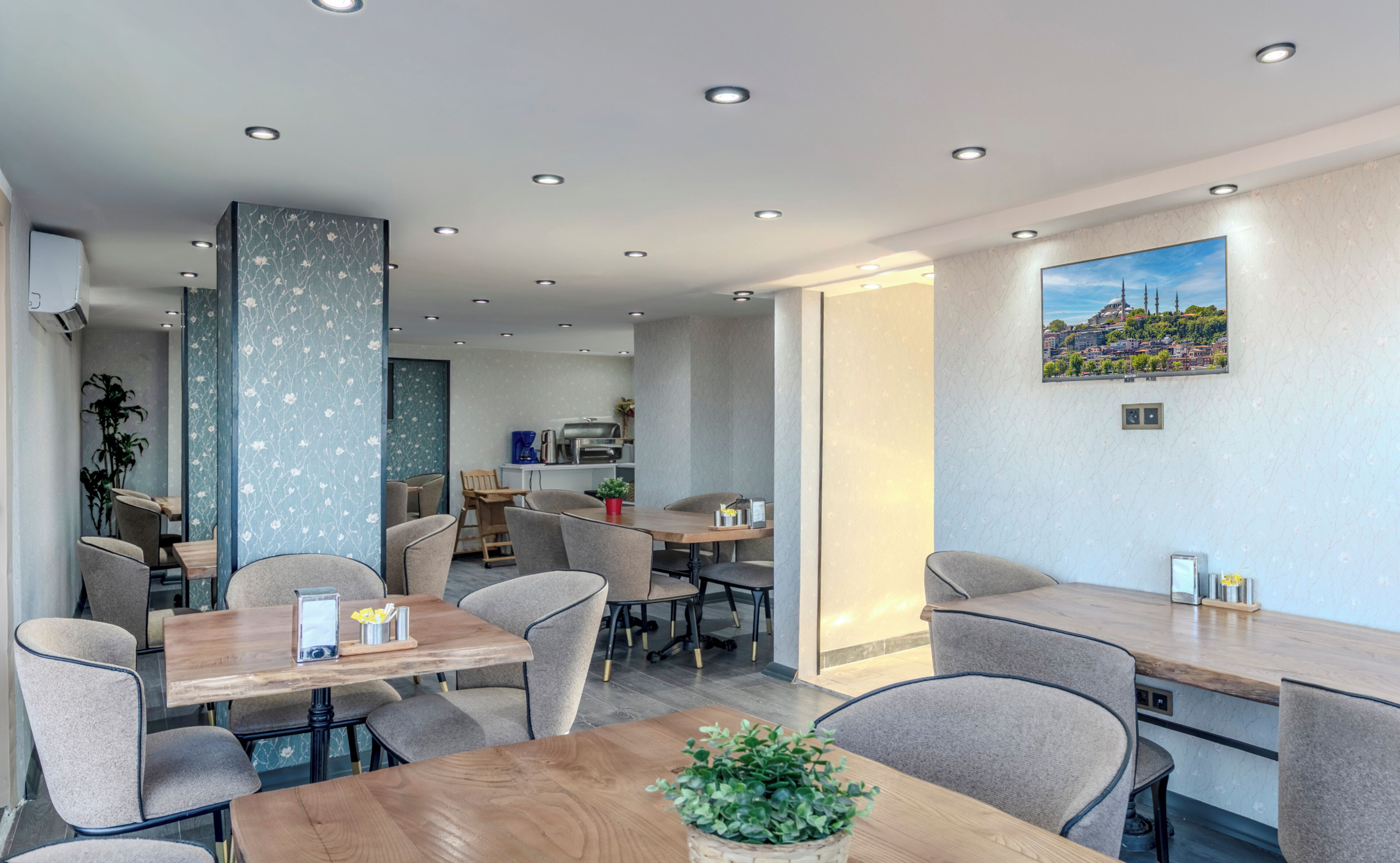 A modern dining area with multiple tables and chairs, a kitchen in the background, and a cityscape painting on the wall.