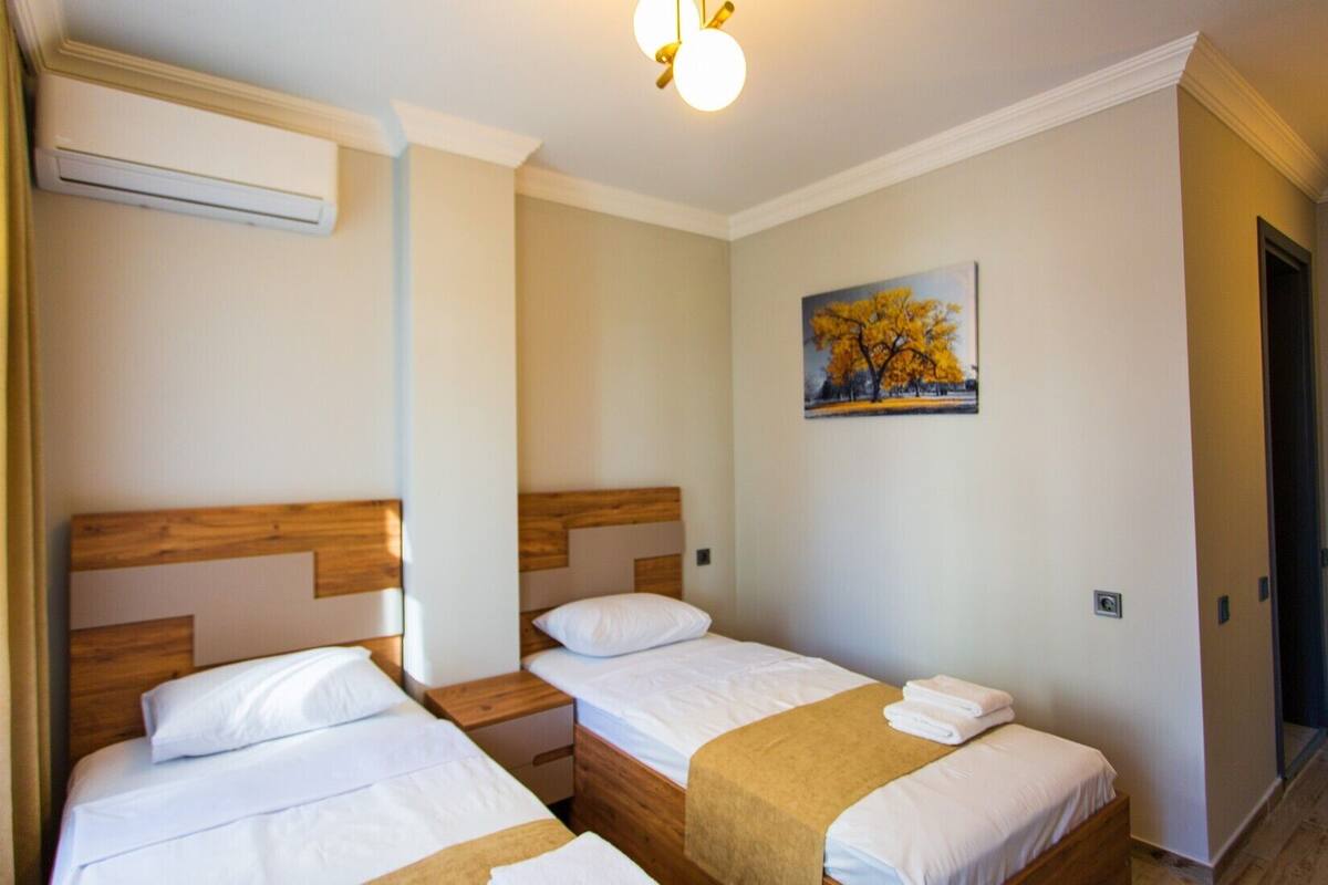 A hotel room with two single beds, wooden headboards, white bedding, a beige throw blanket on one bed, a painting of trees on the wall, and a ceiling light fixture.
