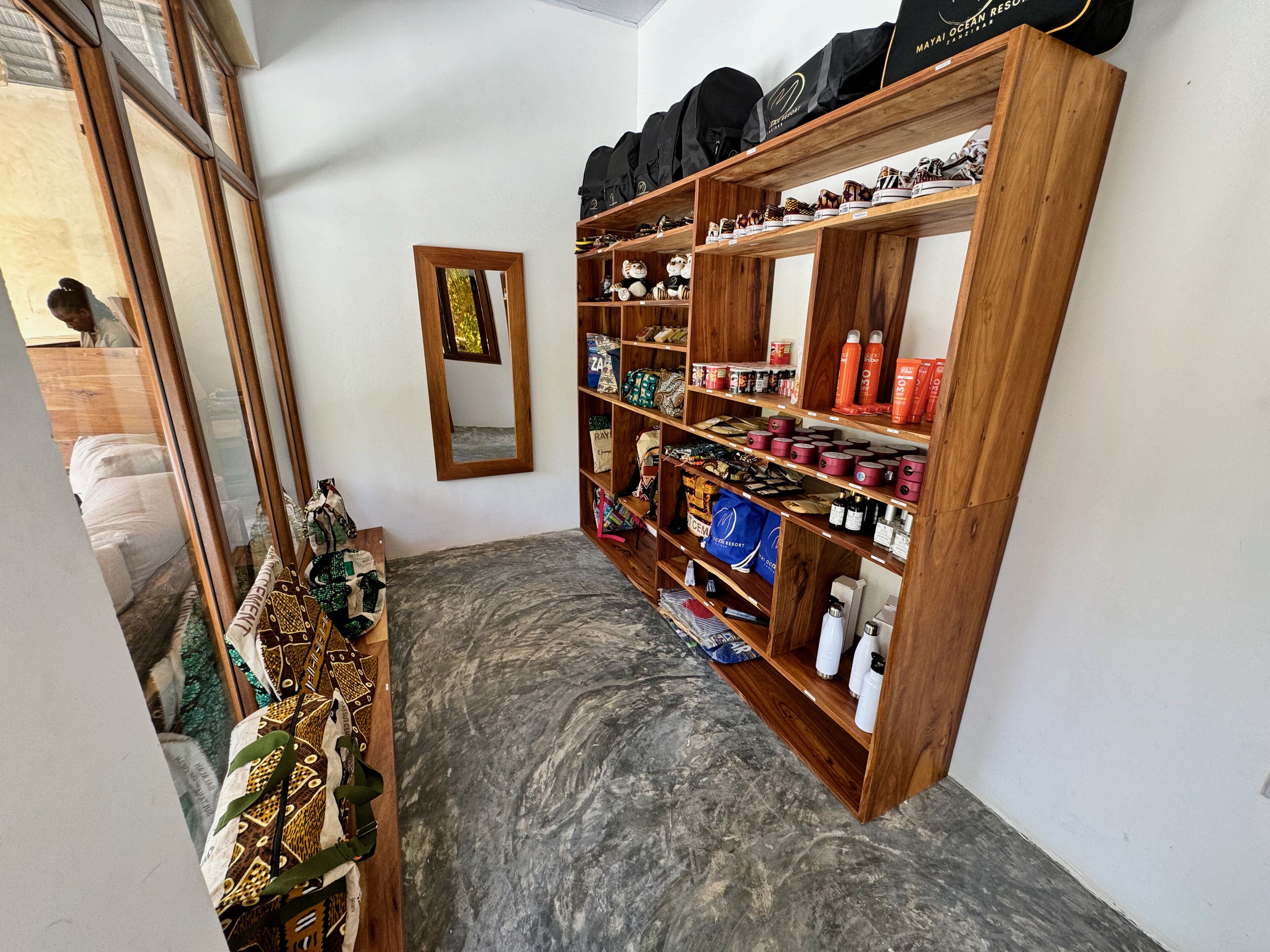 Ein schmaler Flur mit Holzböden, an dem Regale mit verschiedenen Gegenständen stehen. An der Wand hängt ein Spiegel, und im Hintergrund ist ein Bett mit gemusterten Betttextilien zu sehen.