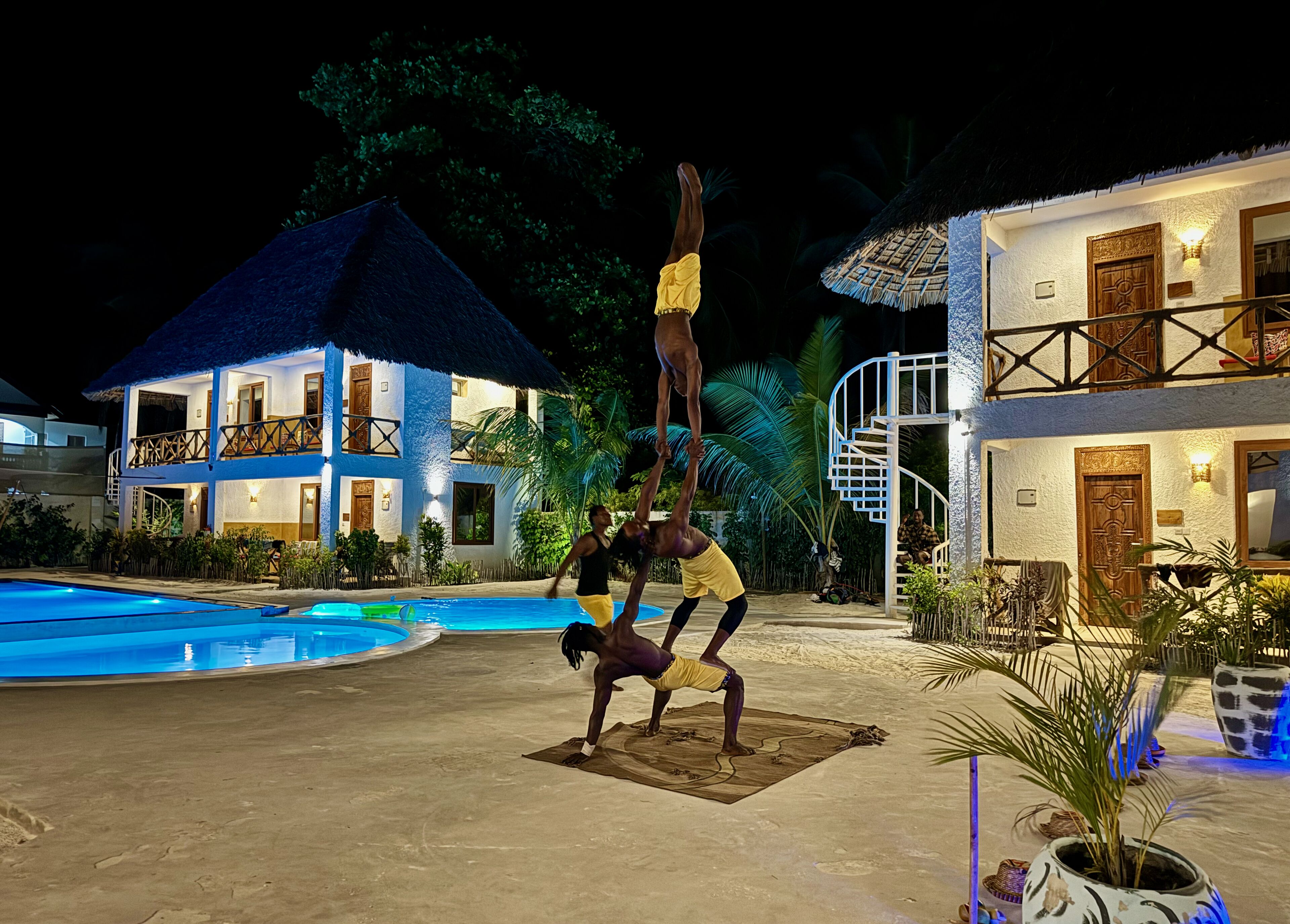 Un hombre realiza una acrobacia sobre una colchoneta cerca de una piscina, con un edificio de resort al fondo.