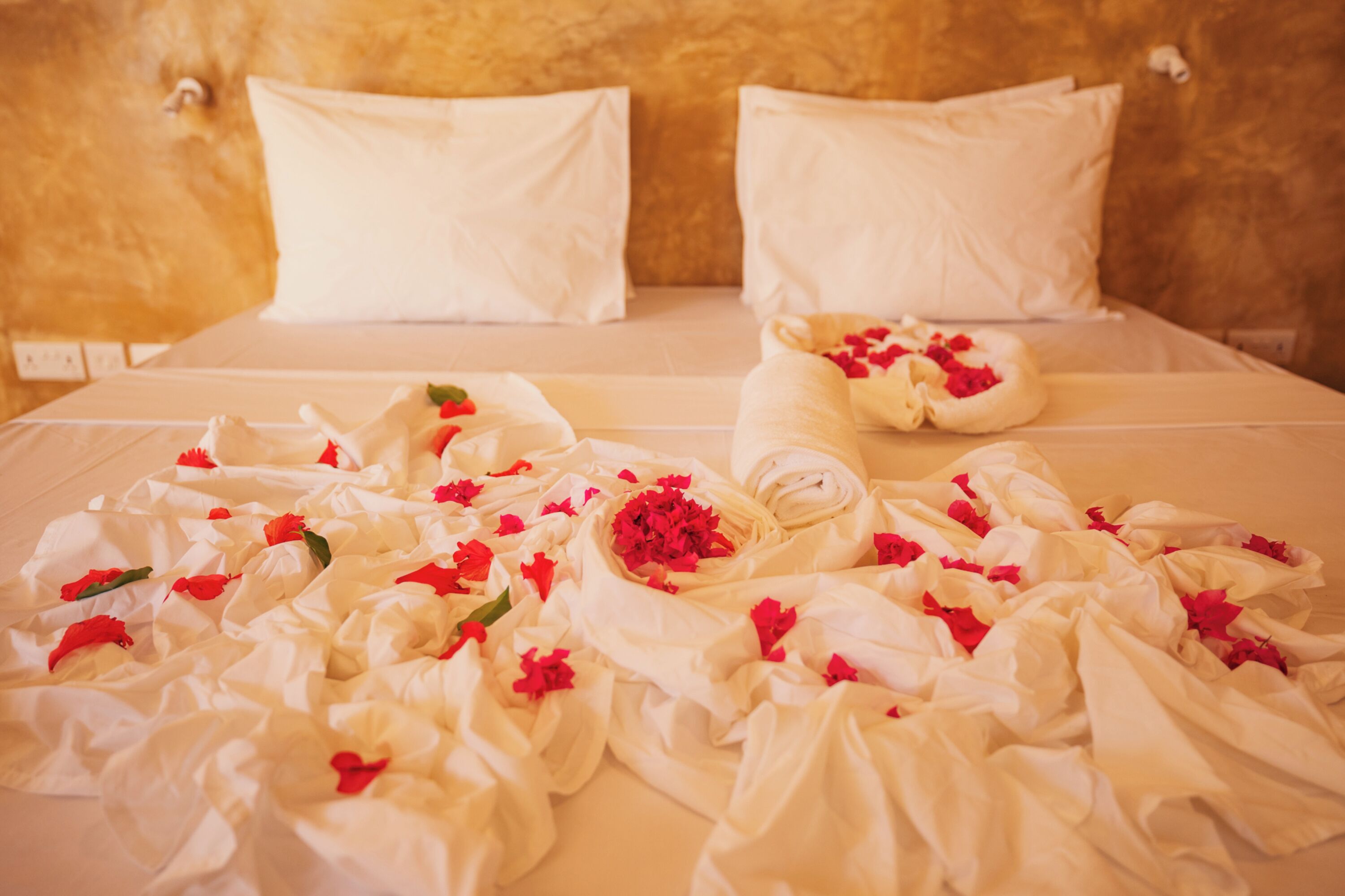 A bed with white linens, pillows, and a rolled towel with red flowers.