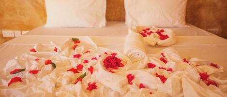 A bed with white linens, pillows, and a rolled towel with red flowers.