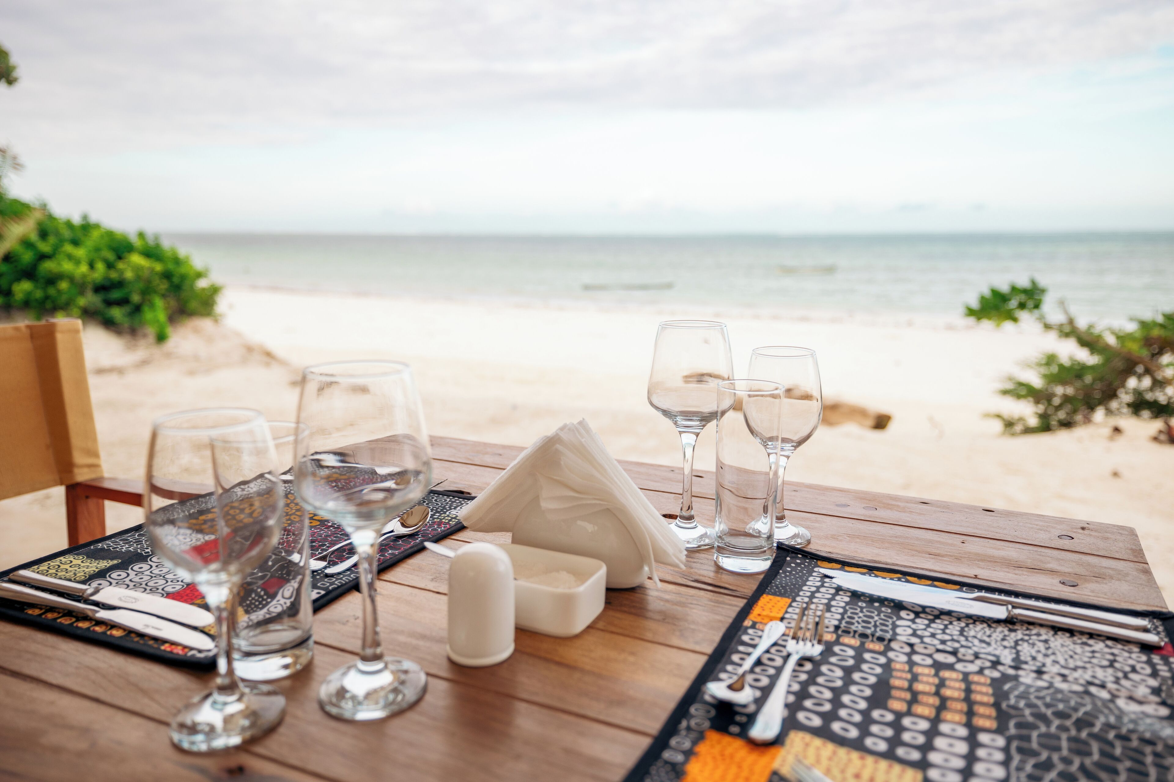 Een eettafel met wijnglazen, bestek en een servet, met uitzicht op een strand.