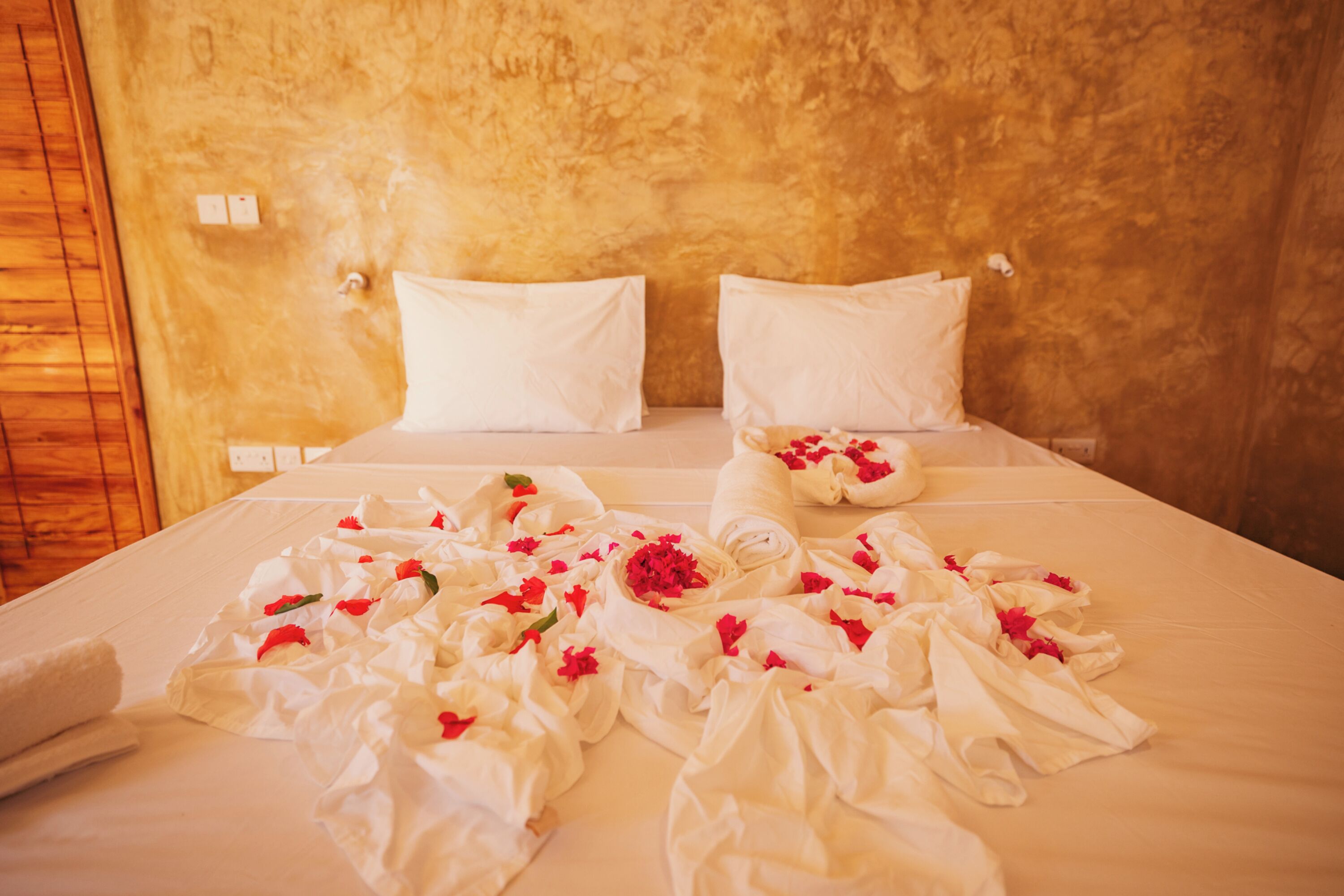 Una cama bien hecha con sábanas blancas y flores rojas, frente a una pared con textura.