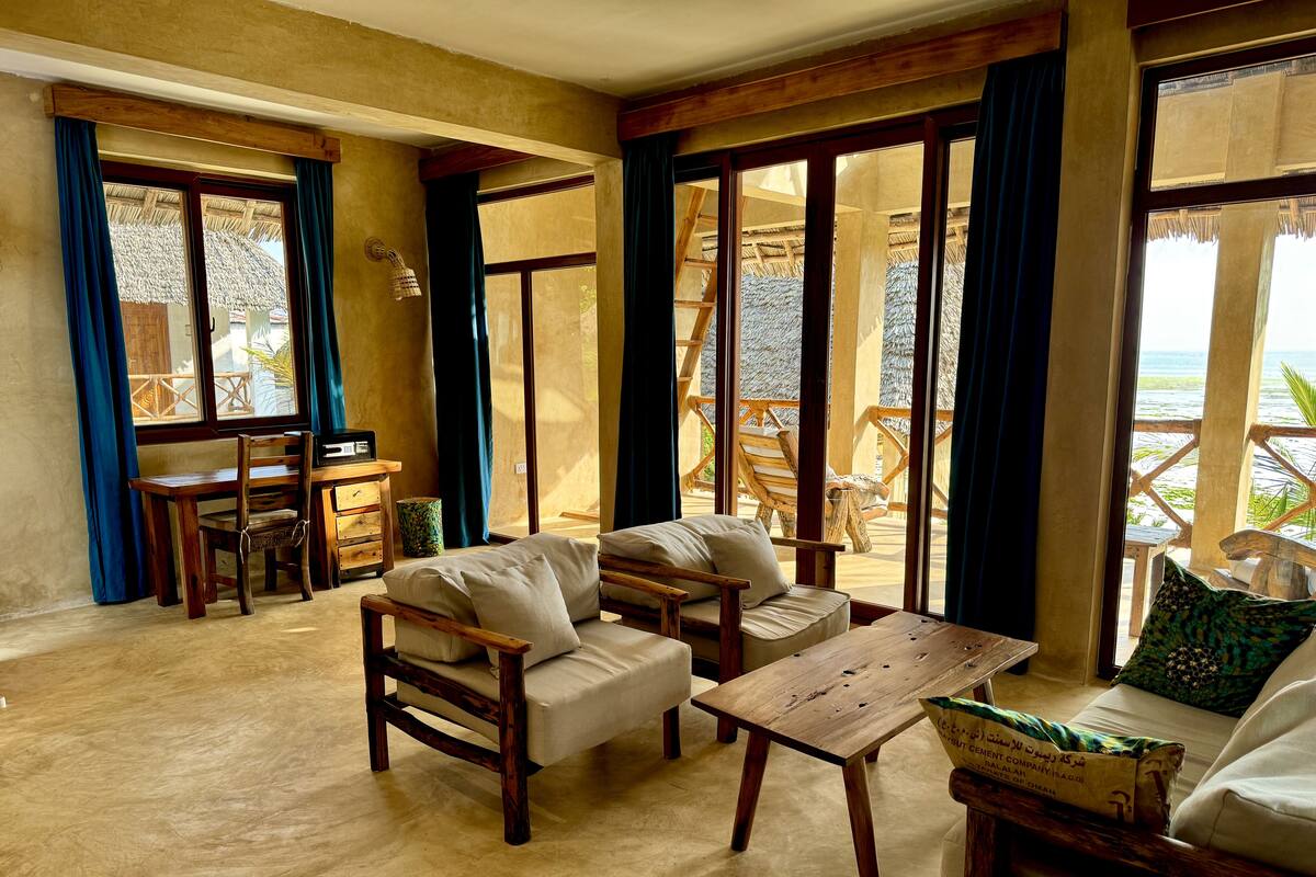 Una habitación con un sofá y una mesa de madera, además de una silla. Hay una ventana con cortinas, y se puede ver una vista de la playa a través de la ventana.