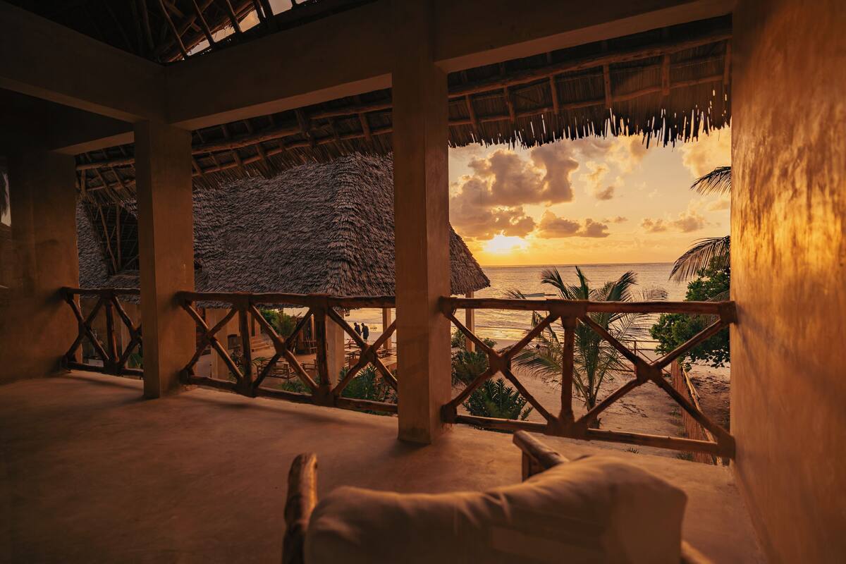 Una habitación frente a la playa con techo de paja, barandillas de madera y vista al atardecer sobre el océano.