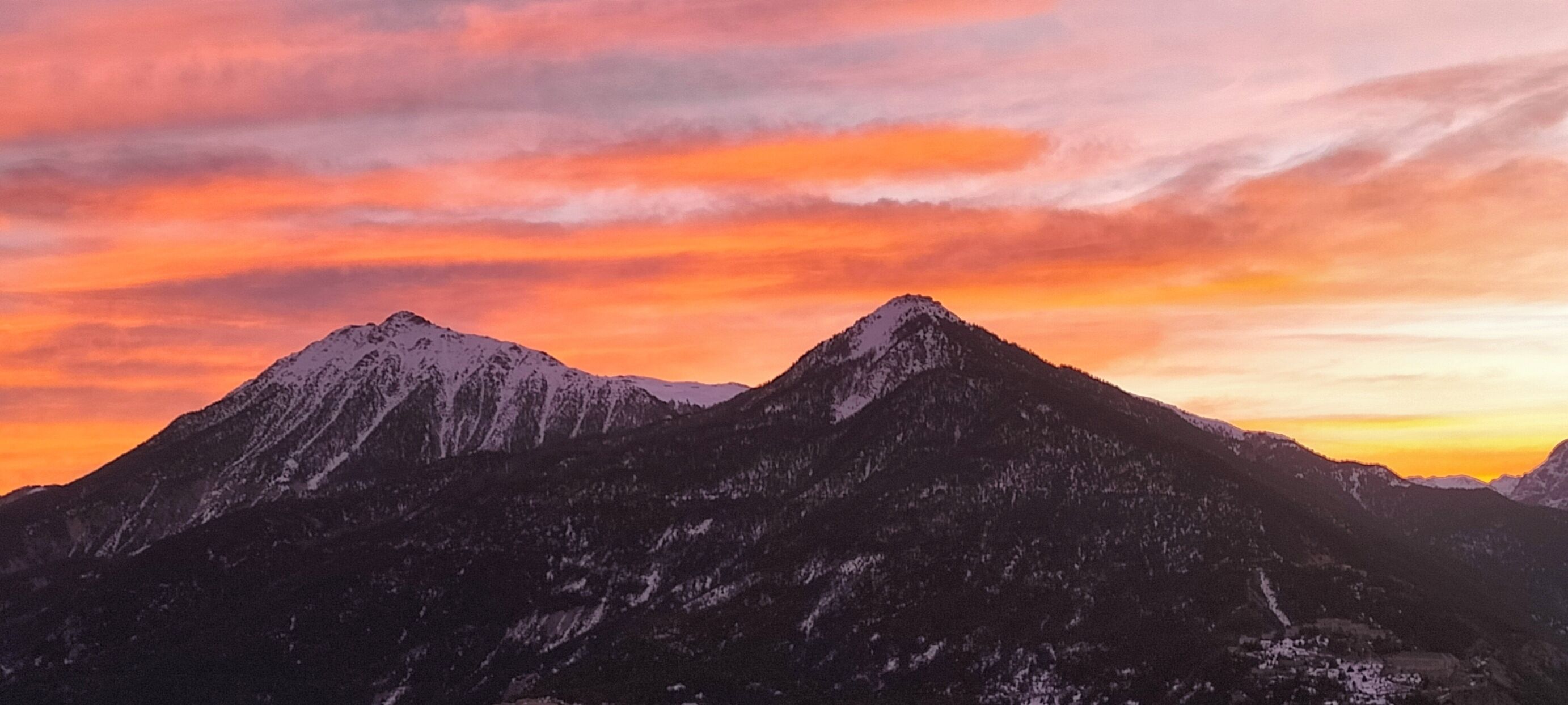 Une chaîne de montagnes aux sommets enneigés, sous un ciel embrasé par le coucher du soleil.