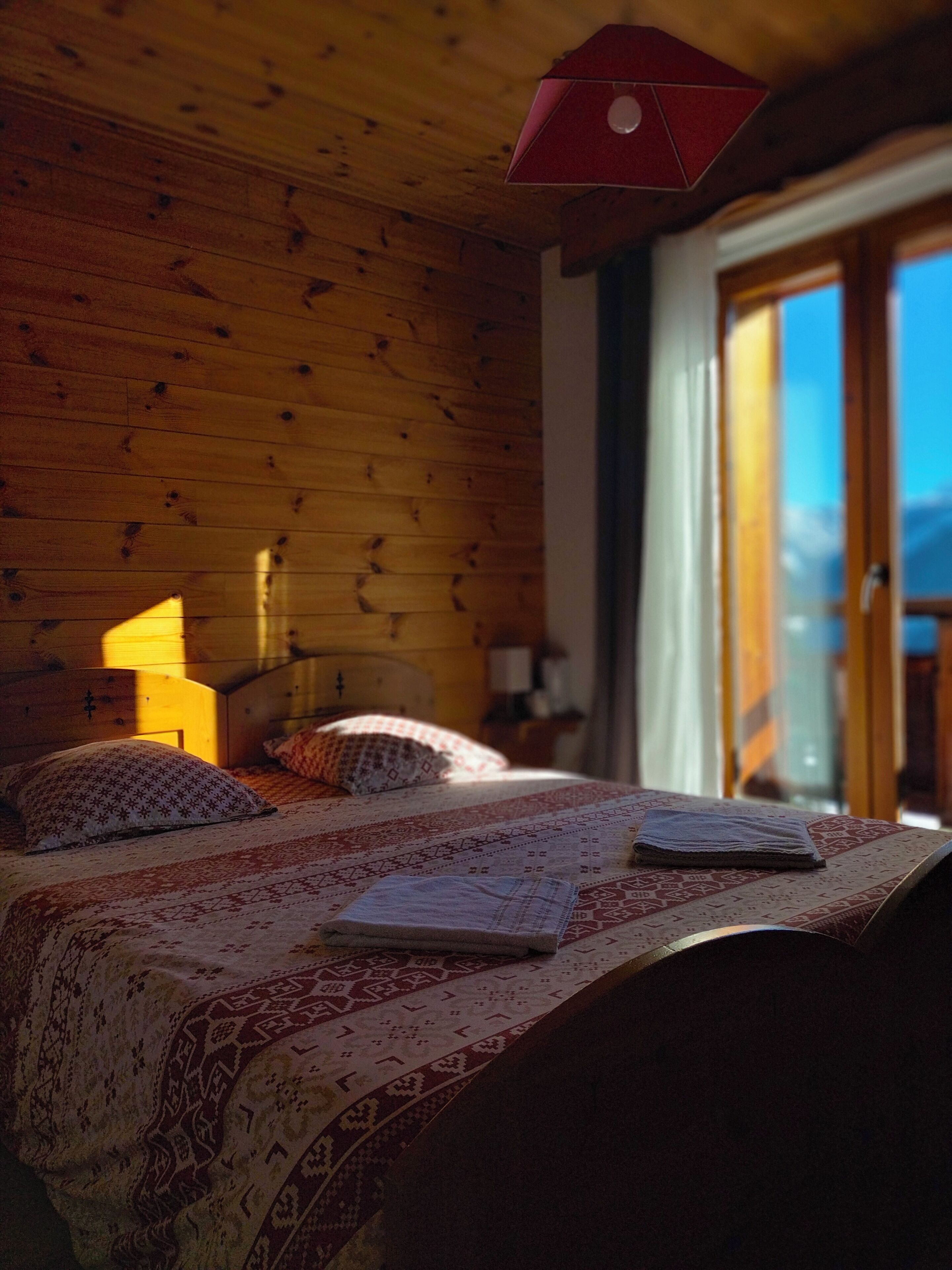Camera da letto in una baita di legno, con un letto, cuscini e una lampada rossa.