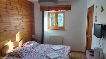 Ein Schlafzimmer mit einem Holz-Kopfteil, einem Bett mit Blumenmuster-Bettwäsche, einem Fenster mit Aussicht, einem Heizkörper, einem Fernseher und einem Wasserkocher auf einer Holzkommode.