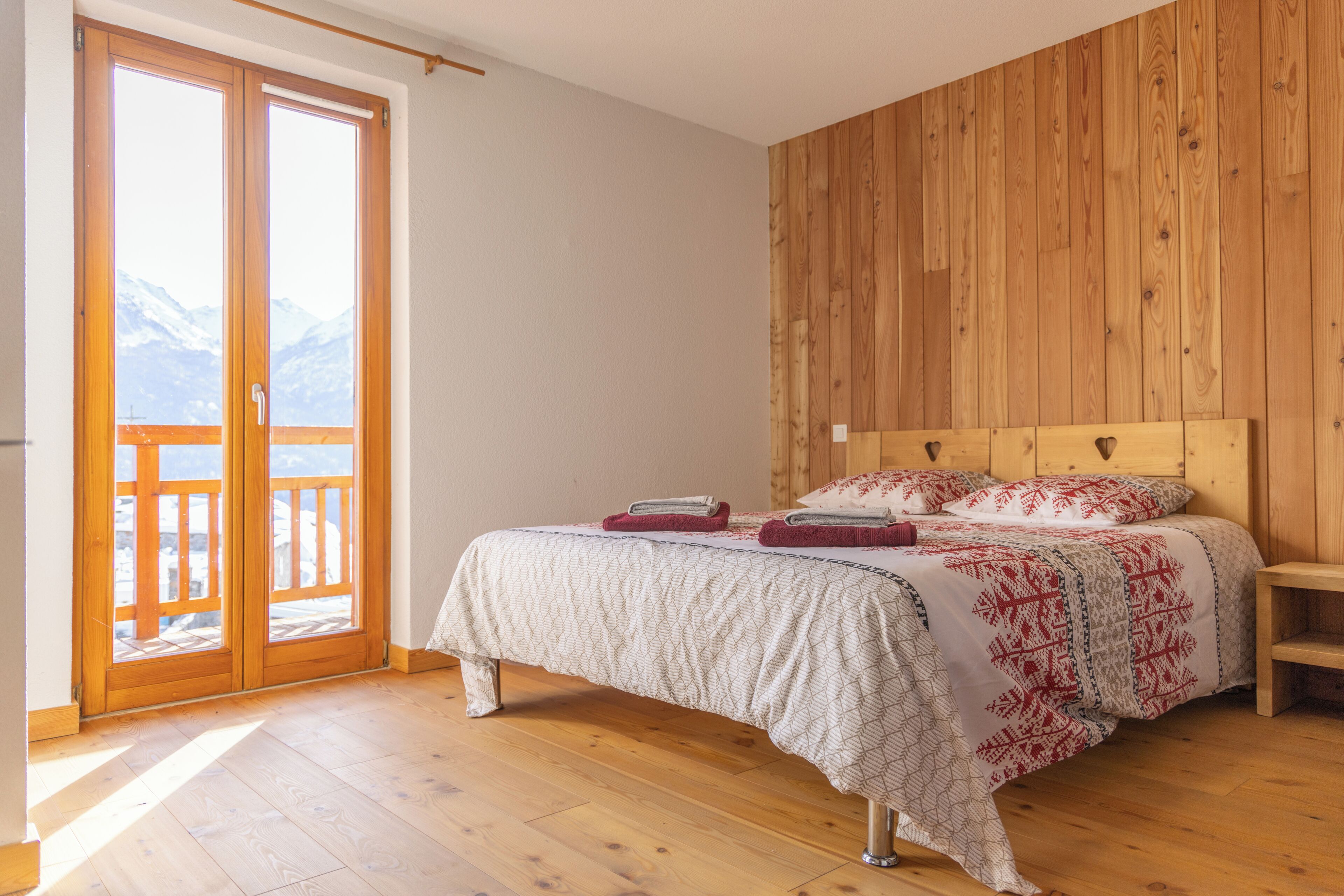 Ein Schlafzimmer mit einem hölzernen Kopfteil, einem Bett mit gemustertem Bettbezug, einem Nachttisch aus Holz und einer Glastür, die auf einen Balkon mit Bergblick führt.