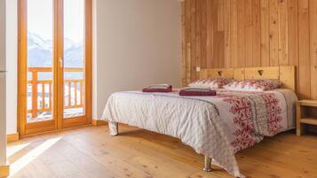 Ein Schlafzimmer mit einem hölzernen Kopfteil, einem Bett mit gemustertem Bettbezug, einem Nachttisch aus Holz und einer Glastür, die auf einen Balkon mit Bergblick führt.