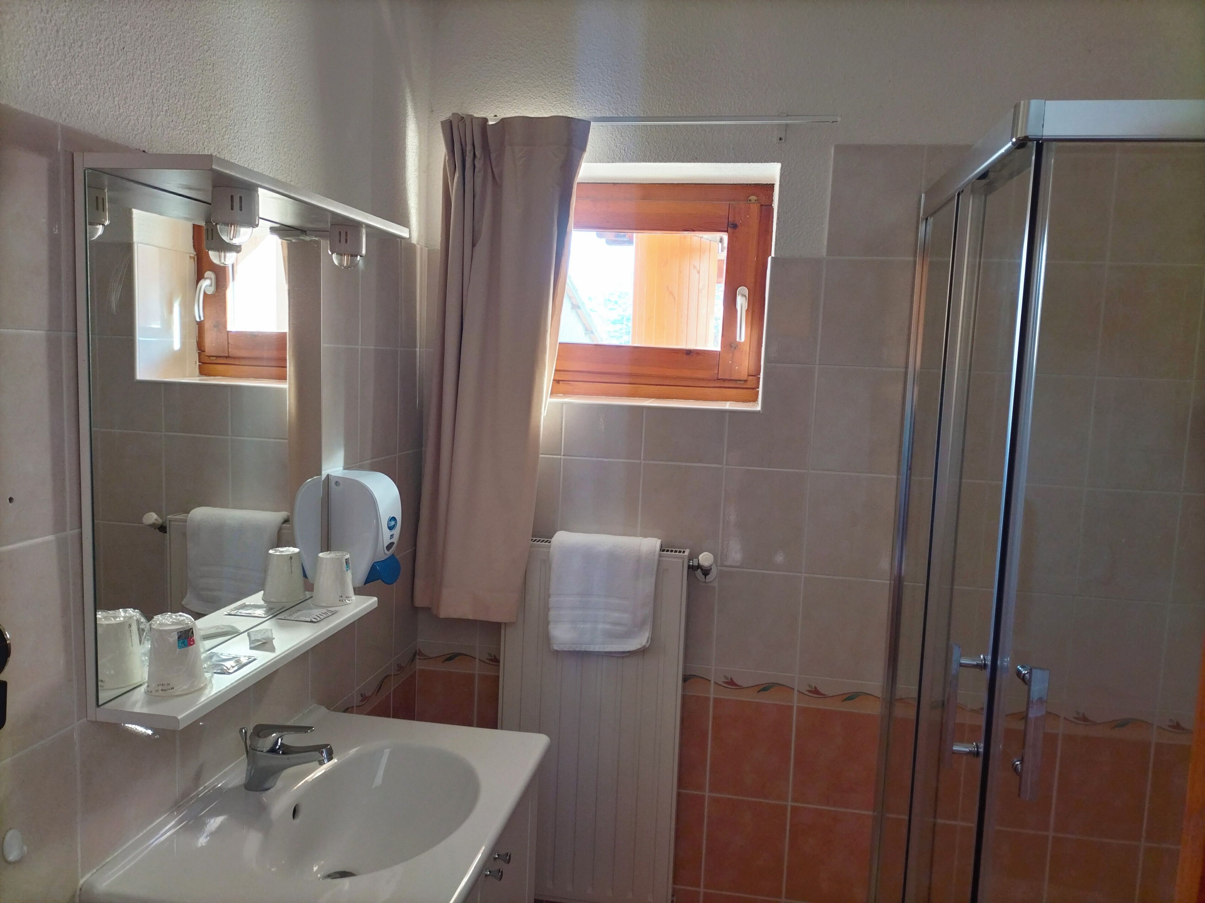 Un baño con lavamanos, espejo, toallero, ducha y ventana con cortinas.