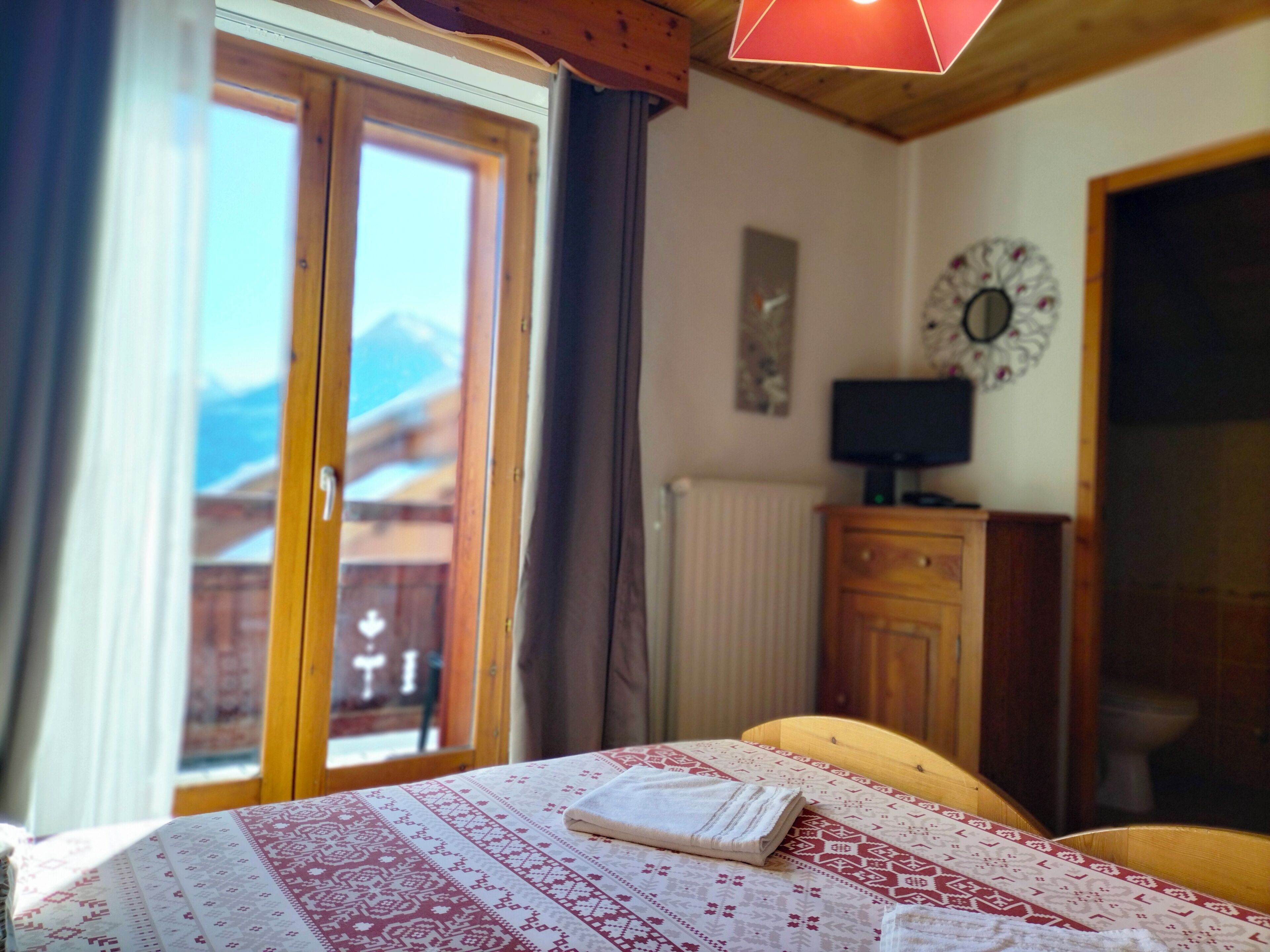 Un dormitorio con cama, televisor sobre un mueble de madera, ventana con vista a las montañas y puerta que da al exterior.
