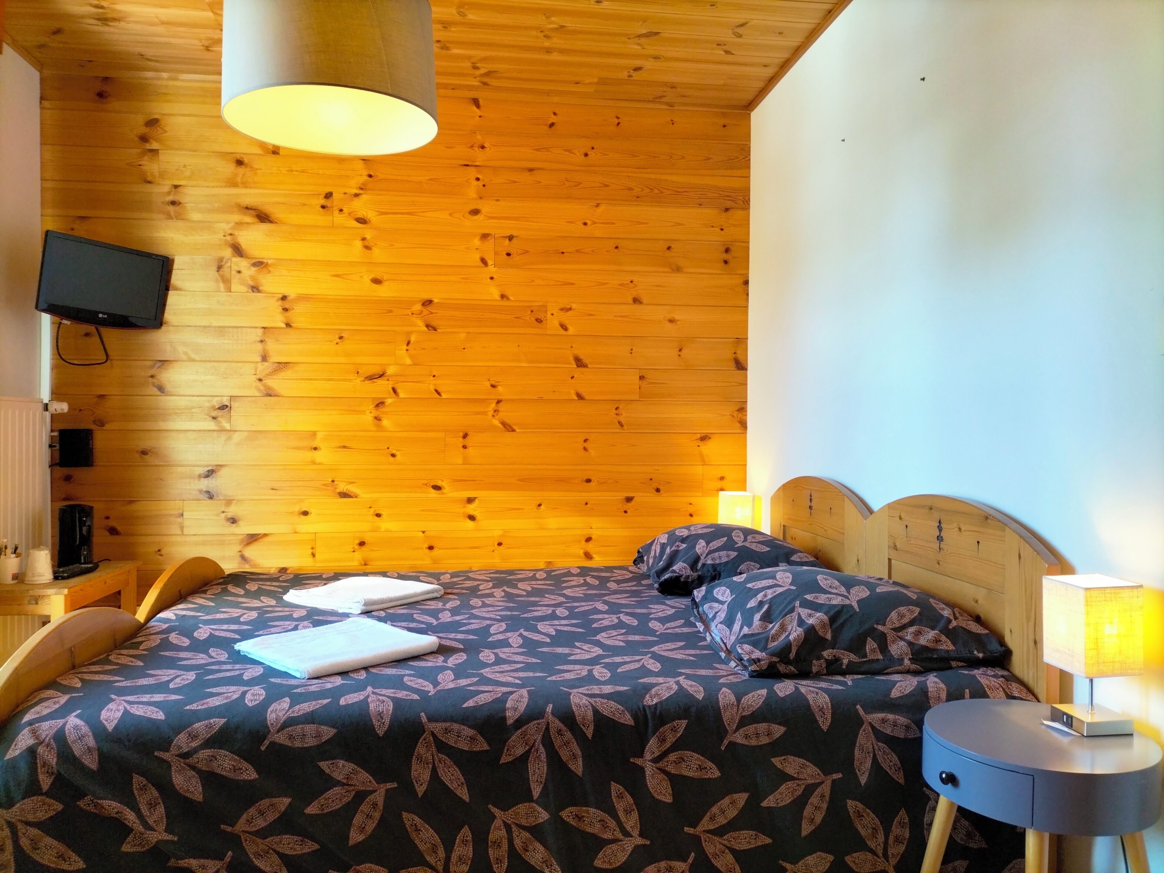 Un dormitorio con techo de madera, una cama con cubrecama estampado con motivos de hojas, una mesita de noche con lámpara y un televisor montado en la pared.