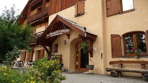 Un edificio de estilo chalete tradicional con un letrero que dice "Maison de Catherine". Tiene balcones de madera, una entrada acogedora con una cesta de flores colgante y mobiliario de exterior.