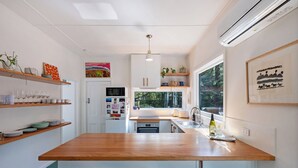 A modern kitchen with a wooden countertop, bar stools, and a view of the outdoors through large windows. - Olive-Nature haven, outdoor bath, artistic cottage, 5 mins to Hyams Beach (Old Erowal Bay)