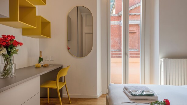 Hotellihuone, jossa on sänky, työpöytä, tuoli, peili ja ikkuna, josta on näkymä punaiseksi maalattuun tiilitaloon.