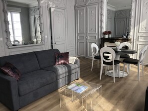 Una sala de estar con un sofá gris, sillas blancas, una mesa de comedor y una mesa de centro de vidrio transparente.