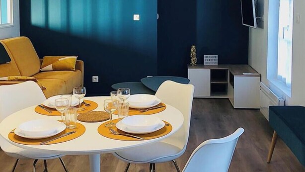 A modern living room with a dark blue accent wall, a sofa, a dining table set for four, and a flat-screen TV mounted on the wall. - Maison Gabriel Appartement Bulle (Abbeville)