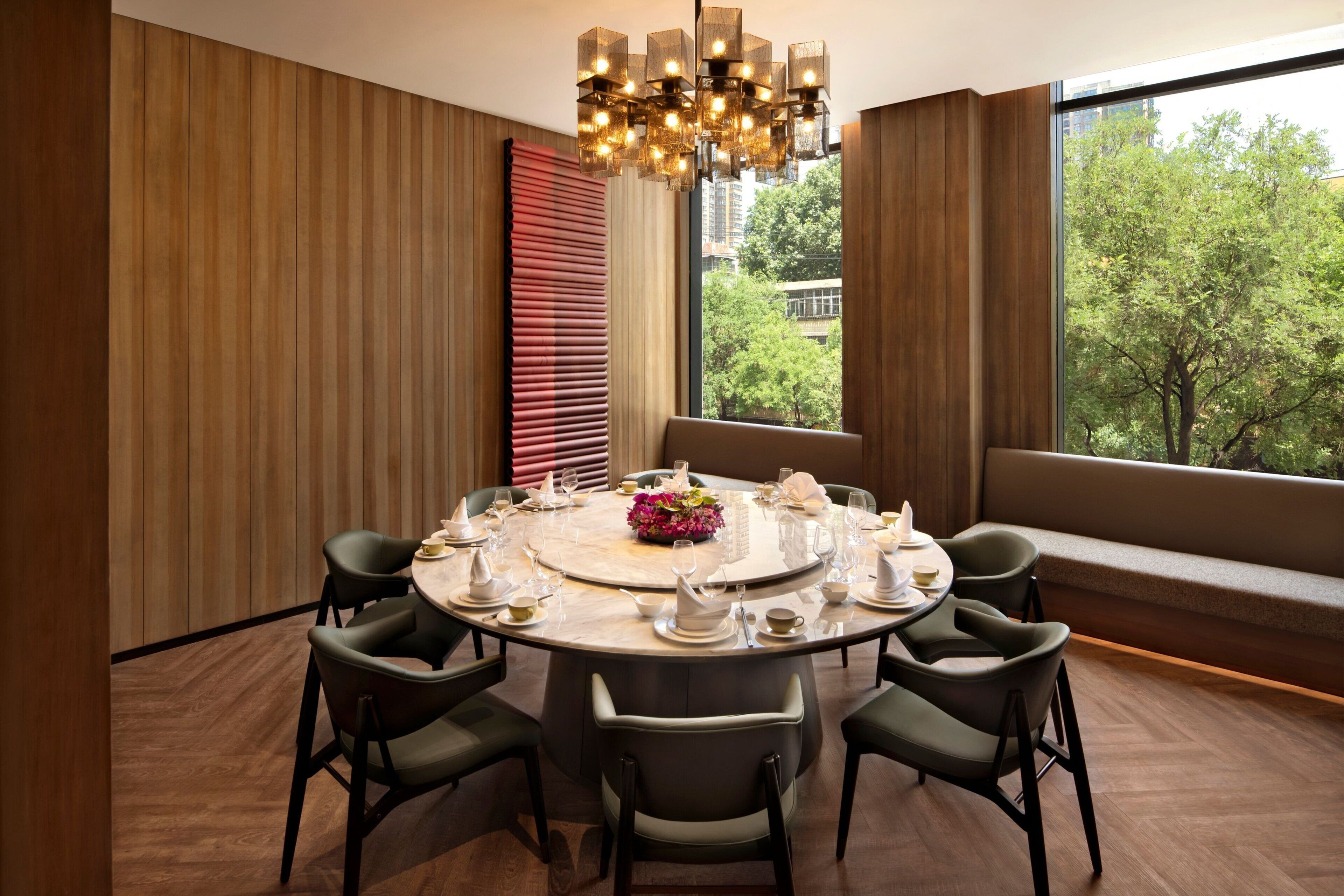Une table de salle à manger ronde, entourée de six chaises, dressée pour un repas, dans une pièce dotée de grandes fenêtres et offrant une vue sur les arbres.