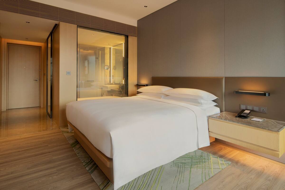 A modern hotel room with a large bed, a bedside table with a phone, and a glass door leading to a bathroom.
