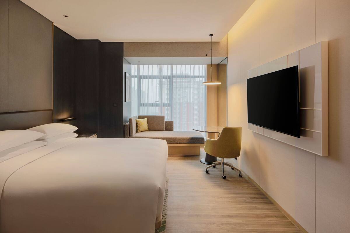 A modern hotel room with a large bed, a desk, a chair, and a TV mounted on the wall.