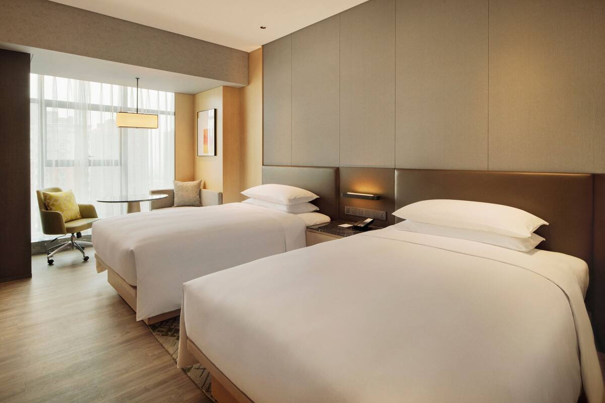 A hotel room with two beds, a desk, a chair, and a large window.