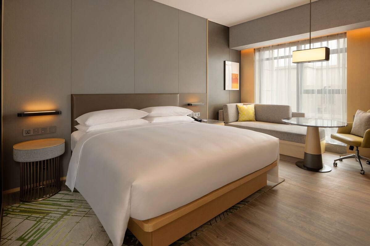 A modern hotel room with a large bed, a round dining table, a sofa, and a small side table.