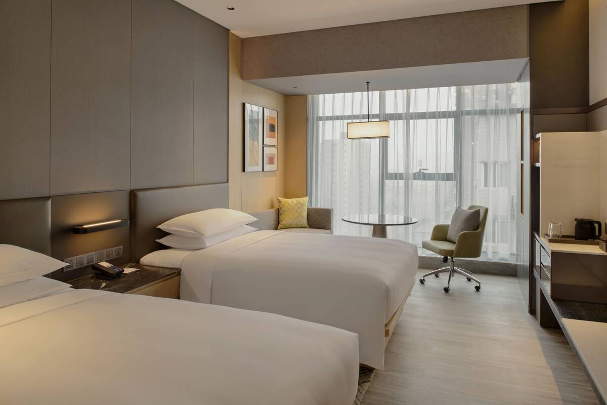 A hotel room with two beds, a small table, a chair, and a view of the city through large windows.