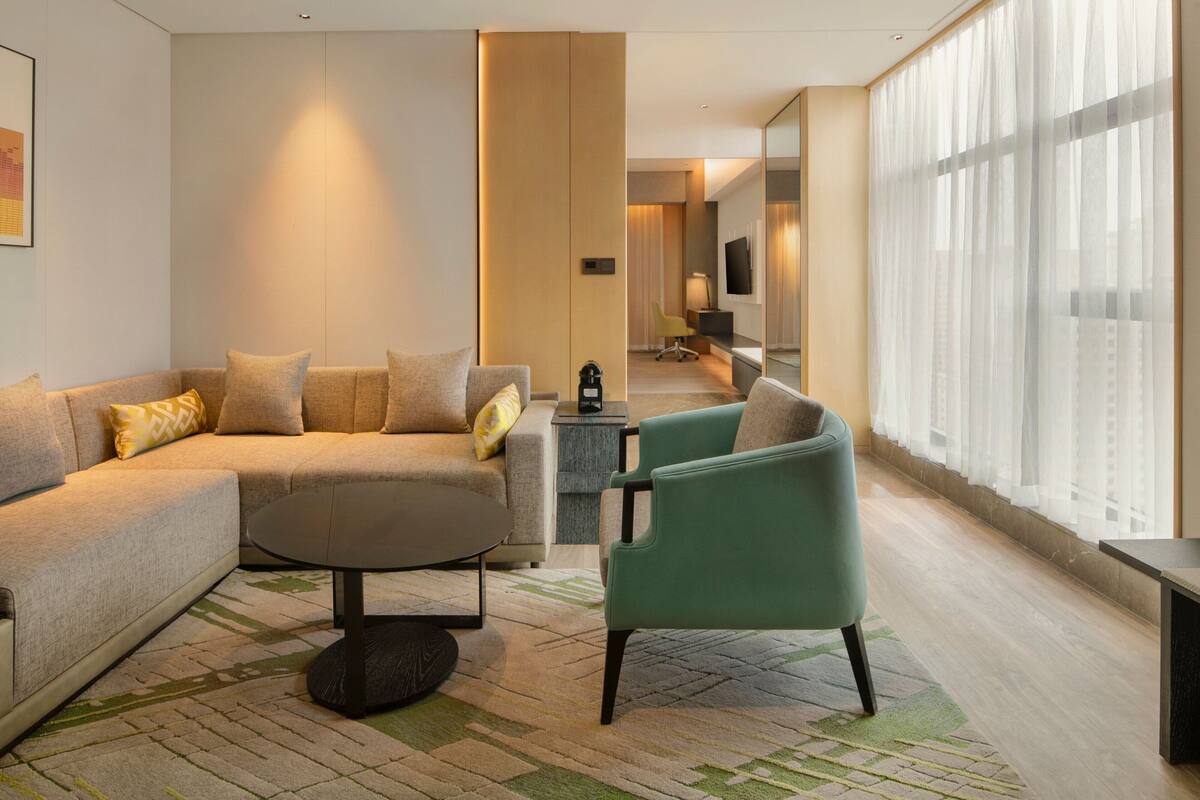 A modern hotel room with a sectional sofa, a round coffee table, a green armchair, and a large window with sheer curtains.