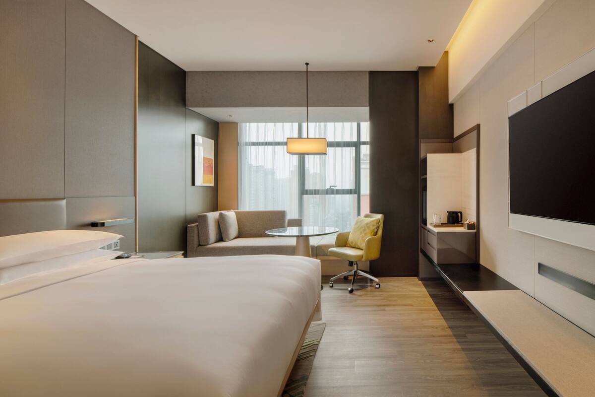 A modern hotel room with a large bed, a seating area with a chair and sofa, a small round table, and a TV mounted on the wall.