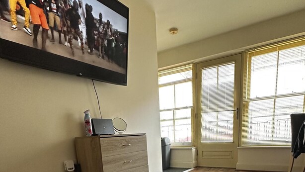 Ein geräumiges Wohnzimmer mit einem großen schwarz-weiß gemusterten Teppich, einem Holzschrank, einem an der Wand befestigten Fernseher und einem großen Fenster mit Jalousien.