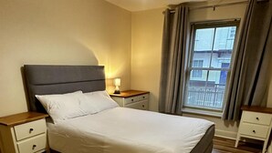 Ein Schlafzimmer mit einem großen Bett, Holzboden, einem Nachttisch, einer Nachttischlampe und einem Fenster mit Vorhängen.